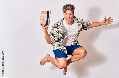 Young handsome hispanic tourist man on vacation wearing summer clothes smiling happy. Jumping with smile on face holding hat over isolated white background