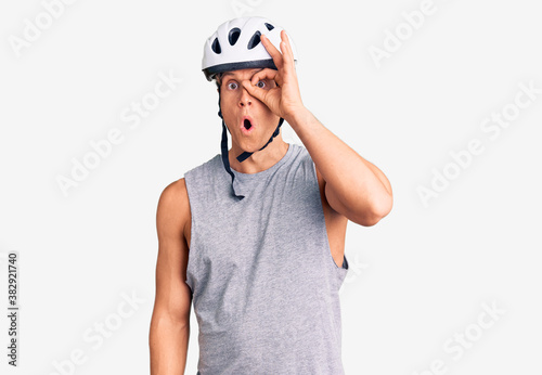 Young handsome man wearing bike helmet doing ok gesture shocked with surprised face, eye looking through fingers. unbelieving expression.