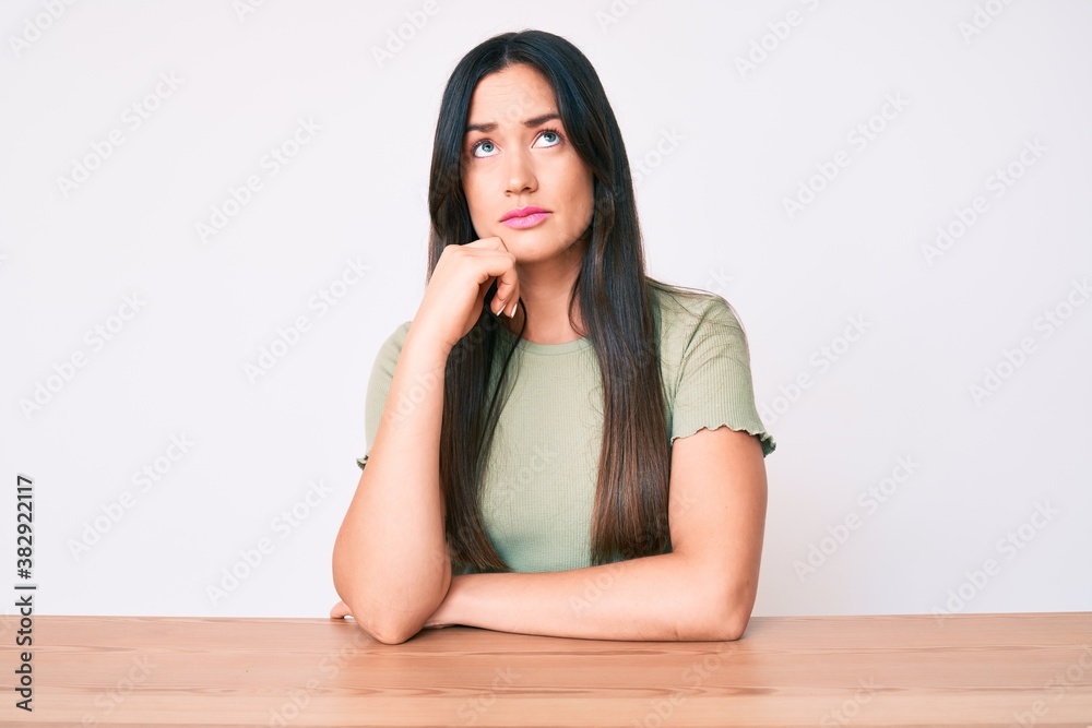 Young caucasian woman sitting at the table wearing casual clothes thinking concentrated about doubt with finger on chin and looking up wondering Young caucasian woman sitting at the table wearing casual clothes thinking concentrated about doubt with finger on chin and looking up wondering