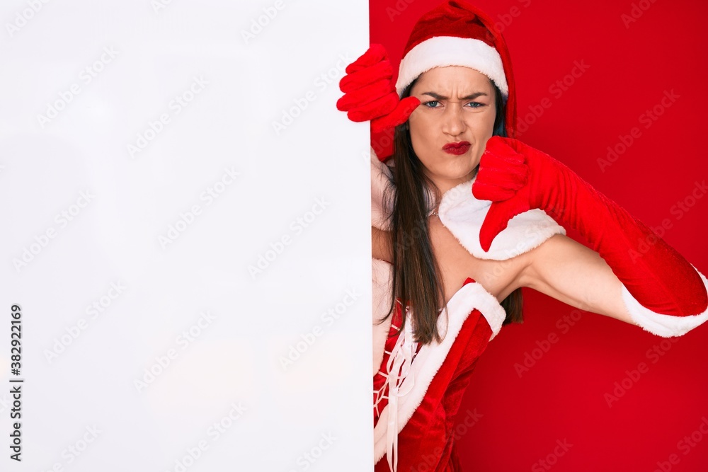 Young woman wearing santa claus costume holding blank empty banner with angry face, negative sign showing dislike with thumbs down, rejection concept Young woman wearing santa claus costume holding blank empty banner with angry face, negative sign showing dislike with thumbs down, rejection concept
