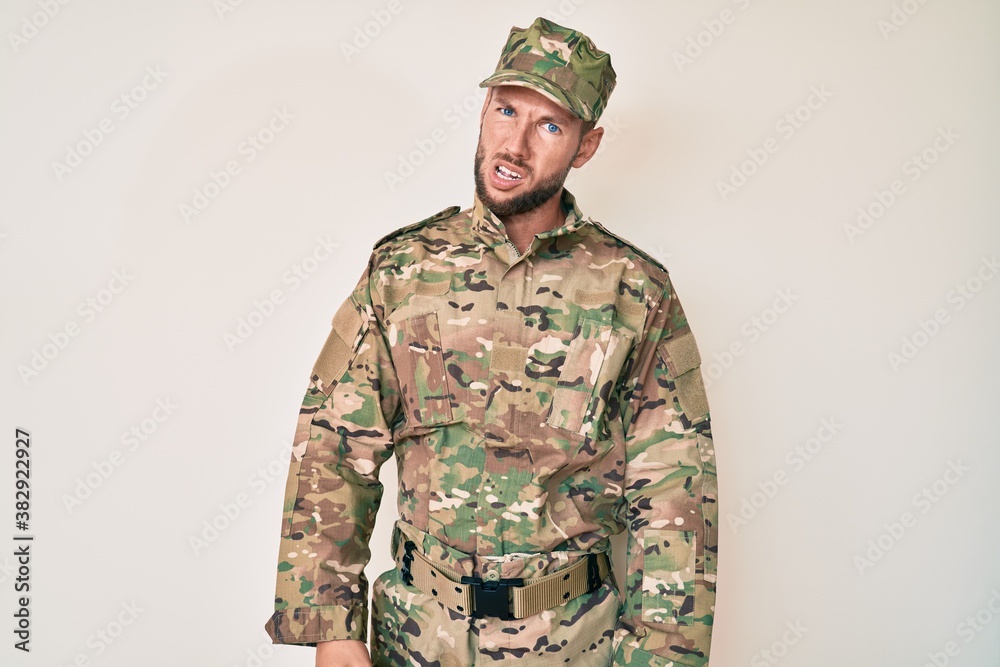 Young caucasian man wearing camouflage army uniform in shock face, looking skeptical and sarcastic, surprised with open mouth