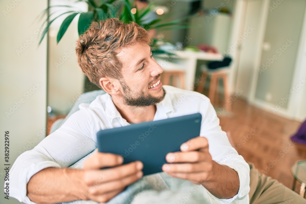 Handsome caucasian man smiling happy sitting on the sofa at home using touchpad device Handsome caucasian man smiling happy sitting on the sofa at home using touchpad device