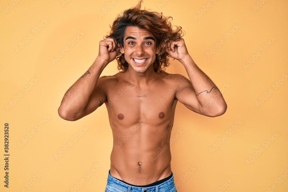 Young hispanic man standing shirtless smiling pulling ears with fingers, funny gesture. audition problem