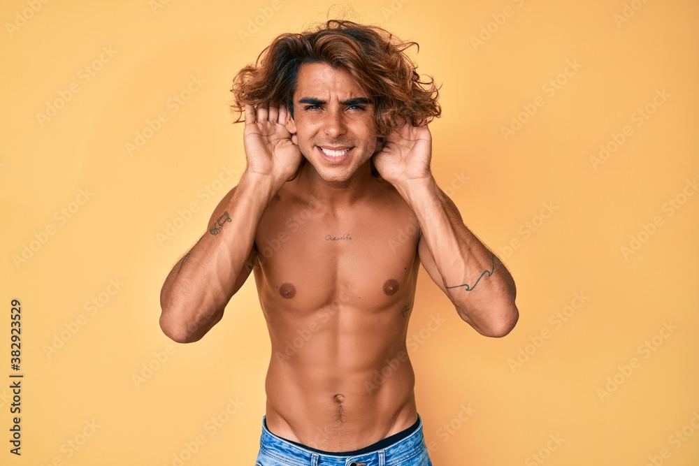 Fototapeta premium Young hispanic man standing shirtless trying to hear both hands on ear gesture, curious for gossip. hearing problem, deaf