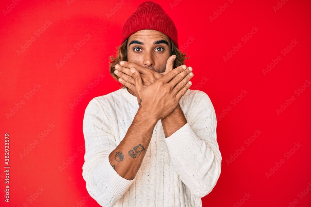 Young hispanic man wearing wool sweater and winter hat shocked covering mouth with hands for mistake. secret concept. Young hispanic man wearing wool sweater and winter hat shocked covering mouth with hands for mistake. secret concept.