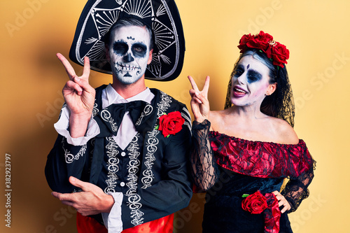 Young couple wearing mexican day of the dead costume over yellow smiling with happy face winking at the camera doing victory sign with fingers. number two.