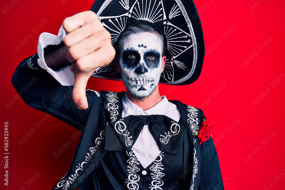 Young man wearing mexican day of the dead costume over red looking unhappy and angry showing rejection and negative with thumbs down gesture. bad expression.