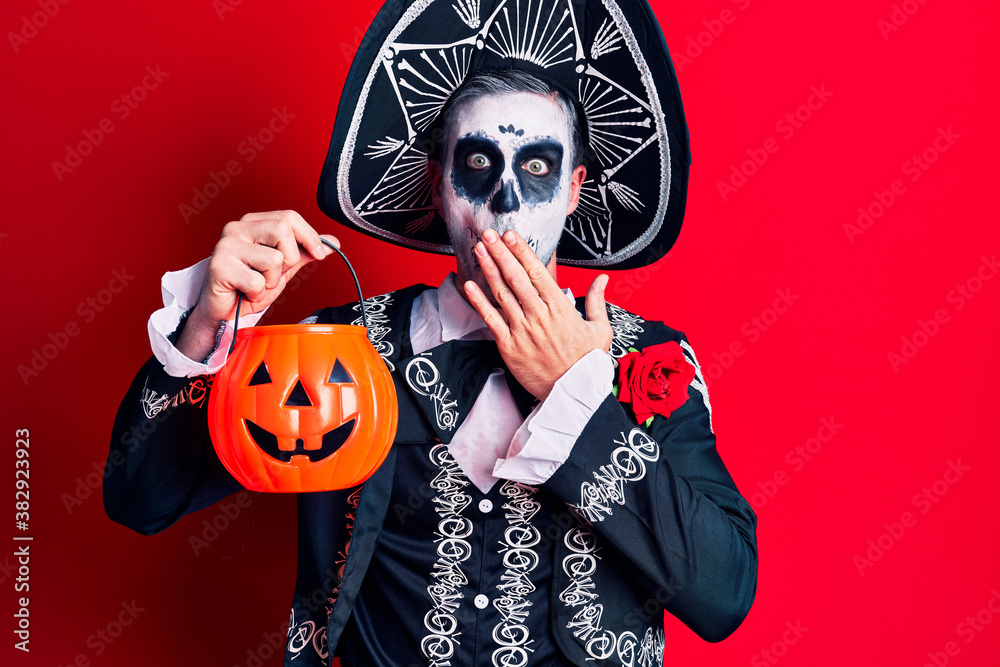 Young man wearing mexican day of the dead costume holding pumpkin covering mouth with hand, shocked and afraid for mistake. surprised expression