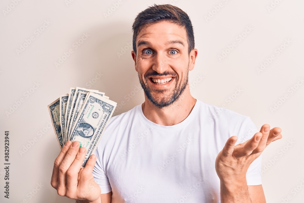 Young handsome man holding bunch of dollars banknotes over isolated white background celebrating achievement with happy smile and winner expression with raised hand Young handsome man holding bunch of dollars banknotes over isolated white background celebrating achievement with happy smile and winner expression with raised hand