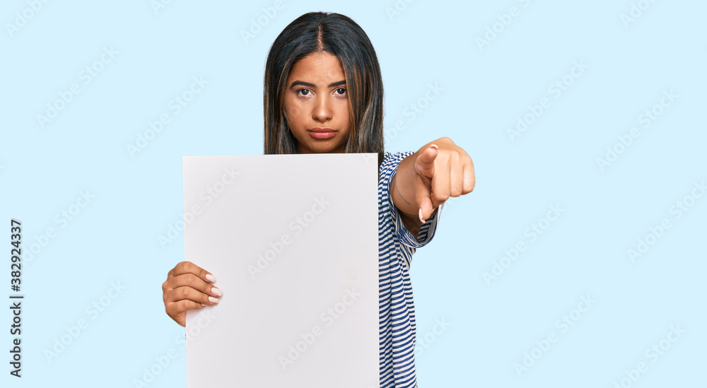 Young latin girl holding blank empty banner pointing with finger to the camera and to you, confident gesture looking serious Young latin girl holding blank empty banner pointing with finger to the camera and to you, confident gesture looking serious