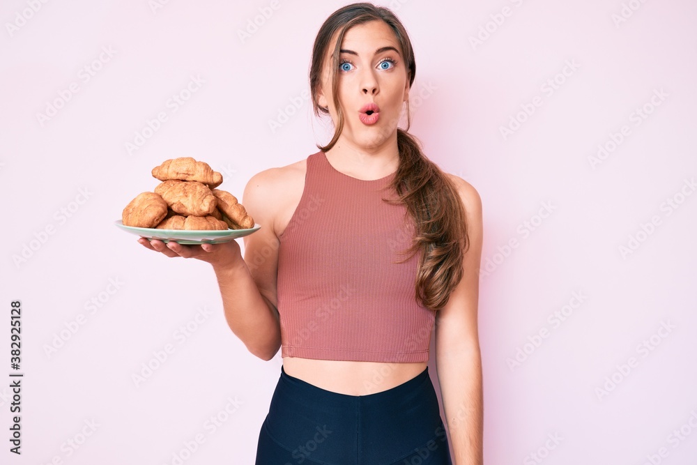 Beautiful young caucasian woman wearing sporty clothes holding croissants scared and amazed with open mouth for surprise, disbelief face Beautiful young caucasian woman wearing sporty clothes holding croissants scared and amazed with open mouth for surprise, disbelief face