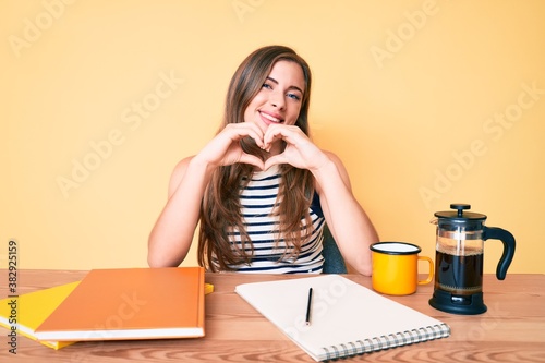 Beautiful young caucasian woman sitting on the table stuying for university smiling in love doing heart symbol shape with hands. romantic concept. photo