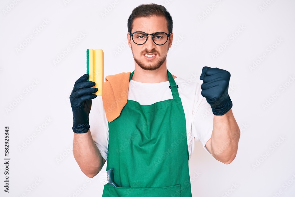 Young handsome man wearing apron holding scourer annoyed and frustrated shouting with anger, yelling crazy with anger and hand raised Young handsome man wearing apron holding scourer annoyed and frustrated shouting with anger, yelling crazy with anger and hand raised