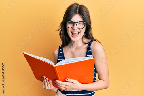 Young beautiful caucasian girl reading a book wearing glasses winking looking at the camera with sexy expression, cheerful and happy face.