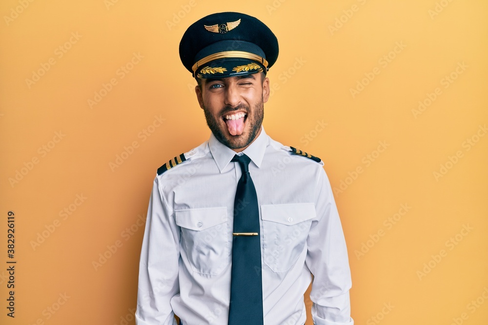 Handsome hispanic man wearing airplane pilot uniform sticking tongue out happy with funny expression. emotion concept. Handsome hispanic man wearing airplane pilot uniform sticking tongue out happy with funny expression. emotion concept.