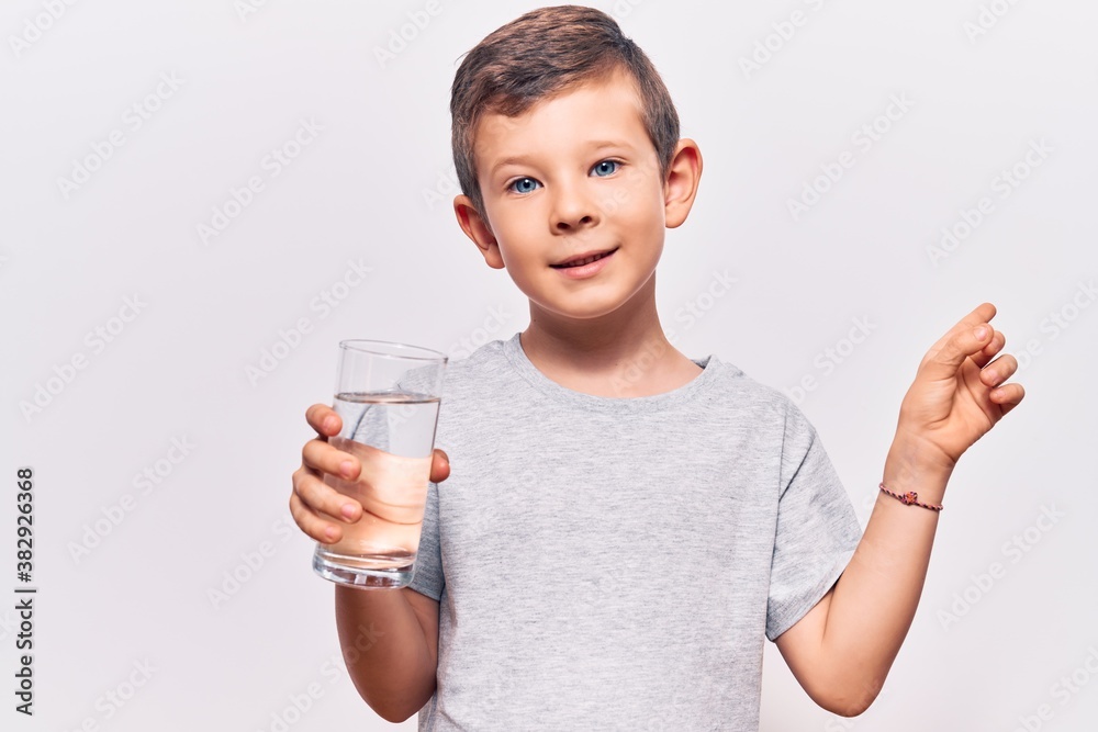 Cute blond kid drinking glass of water smiling happy pointing with hand and finger to the side Cute blond kid drinking glass of water smiling happy pointing with hand and finger to the side