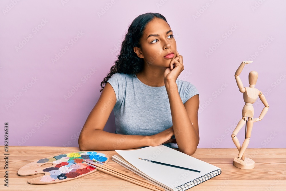 Young african american girl artist sitting at studio table thinking concentrated about doubt with finger on chin and looking up wondering Young african american girl artist sitting at studio table thinking concentrated about doubt with finger on chin and looking up wondering
