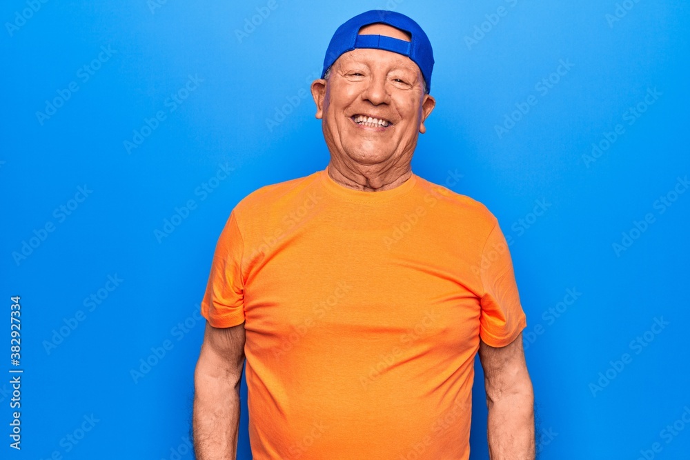 Fototapeta premium Senior handsome grey-haired man wearing casual t-shirt and cap over blue background with a happy and cool smile on face. Lucky person.