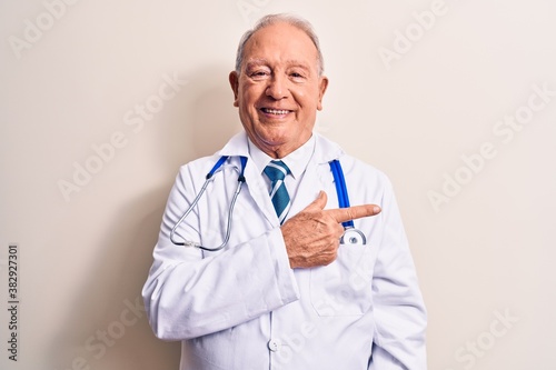 Senior grey-haired doctor man wearing coat and stethoscope standing over white background smiling cheerful pointing with hand and finger up to the side