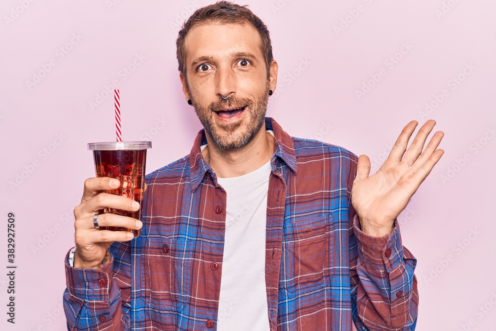 Young handsome man drinking glass of cola beverage celebrating achievement with happy smile and winner expression with raised hand Young handsome man drinking glass of cola beverage celebrating achievement with happy smile and winner expression with raised hand