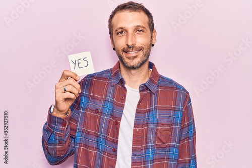 Young handsome man holding yes reminder looking positive and happy standing and smiling with a confident smile showing teeth © Krakenimages.com
