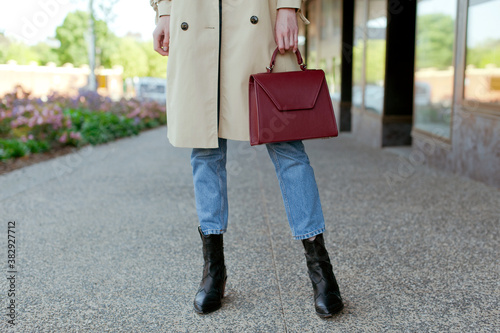 Fashionable young woman wearing beige trench coat, sweater, blue jeans and black high heel cowboy boots. She is holding burgundy colour leather handbag in hand. Street style.