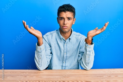 фотографія Young handsome african american man wearing casual clothes sitting on the table clueless and confused with open arms, no idea concept