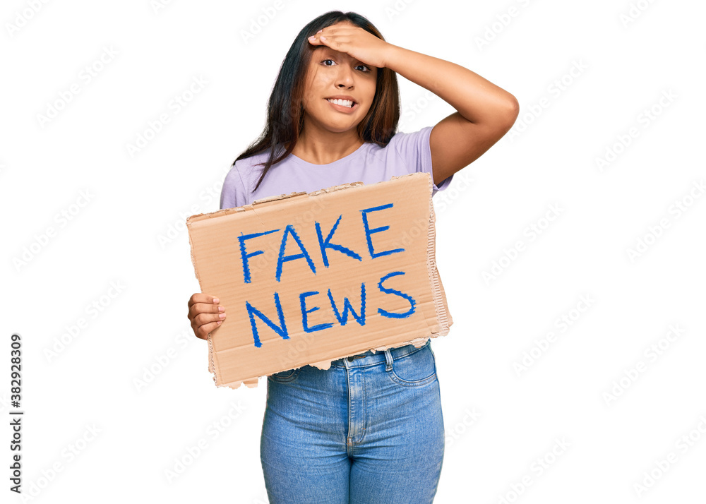 Young latin woman holding fake news banner stressed and frustrated with hand on head, surprised and angry face Young latin woman holding fake news banner stressed and frustrated with hand on head, surprised and angry face