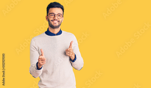 Young handsome hispanic man wearing elegant clothes and glasses pointing fingers to camera with happy and funny face. good energy and vibes.