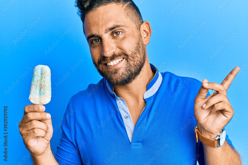 Handsome man with beard holding ice cream smiling happy pointing with hand and finger to the side Handsome man with beard holding ice cream smiling happy pointing with hand and finger to the side