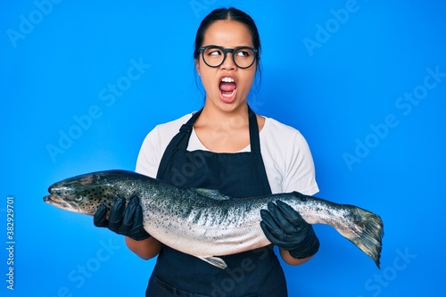 Young beautiful asian girl fishmonger selling fresh raw salmon angry and mad screaming frustrated and furious, shouting with anger. rage and aggressive concept.