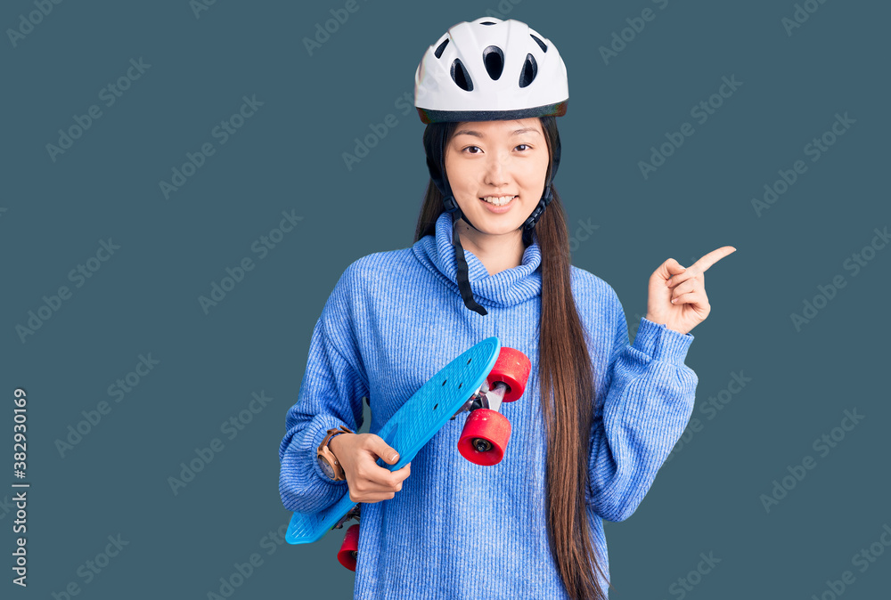 Young beautiful chinese woman wearing security helmet holding skate smiling happy pointing with hand and finger to the side Young beautiful chinese woman wearing security helmet holding skate smiling happy pointing with hand and finger to the side