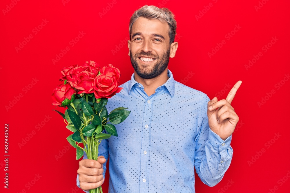 Young handsome blond romantic man holding bouquet of beautiful roses over red background smiling happy pointing with hand and finger to the side Young handsome blond romantic man holding bouquet of beautiful roses over red background smiling happy pointing with hand and finger to the side