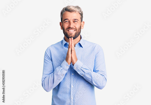 Young handsome blond man wearing elegant shirt praying with hands together asking for forgiveness smiling confident.