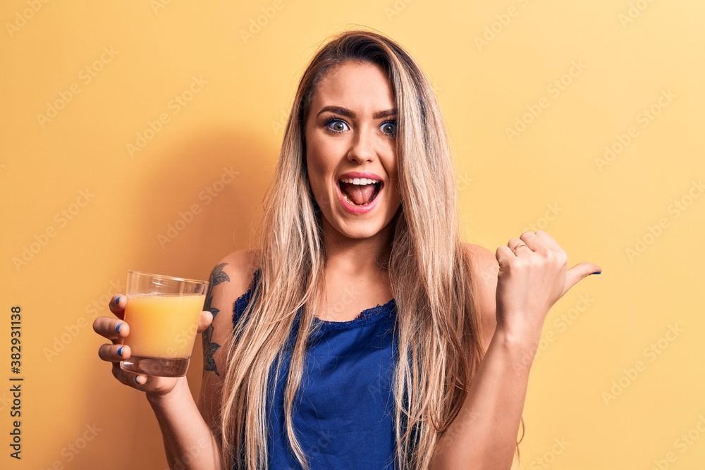Young beautiful blonde woman drinking glass of healthy orange juice over yellow background pointing thumb up to the side smiling happy with open mouth Young beautiful blonde woman drinking glass of healthy orange juice over yellow background pointing thumb up to the side smiling happy with open mouth