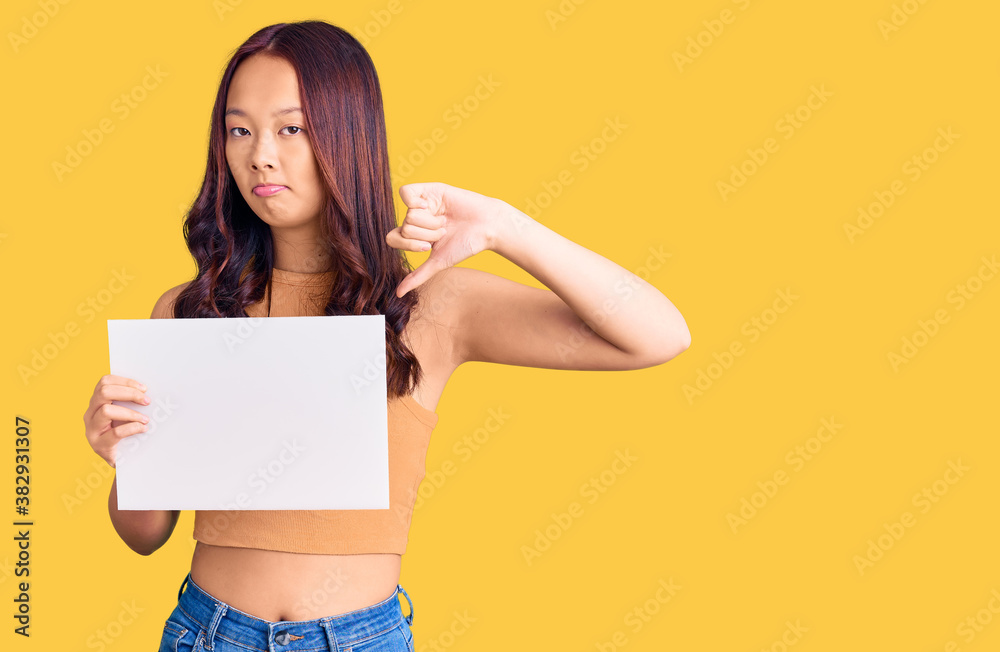 Young beautiful chinese girl holding paper banner with blank space with angry face, negative sign showing dislike with thumbs down, rejection concept Young beautiful chinese girl holding paper banner with blank space with angry face, negative sign showing dislike with thumbs down, rejection concept