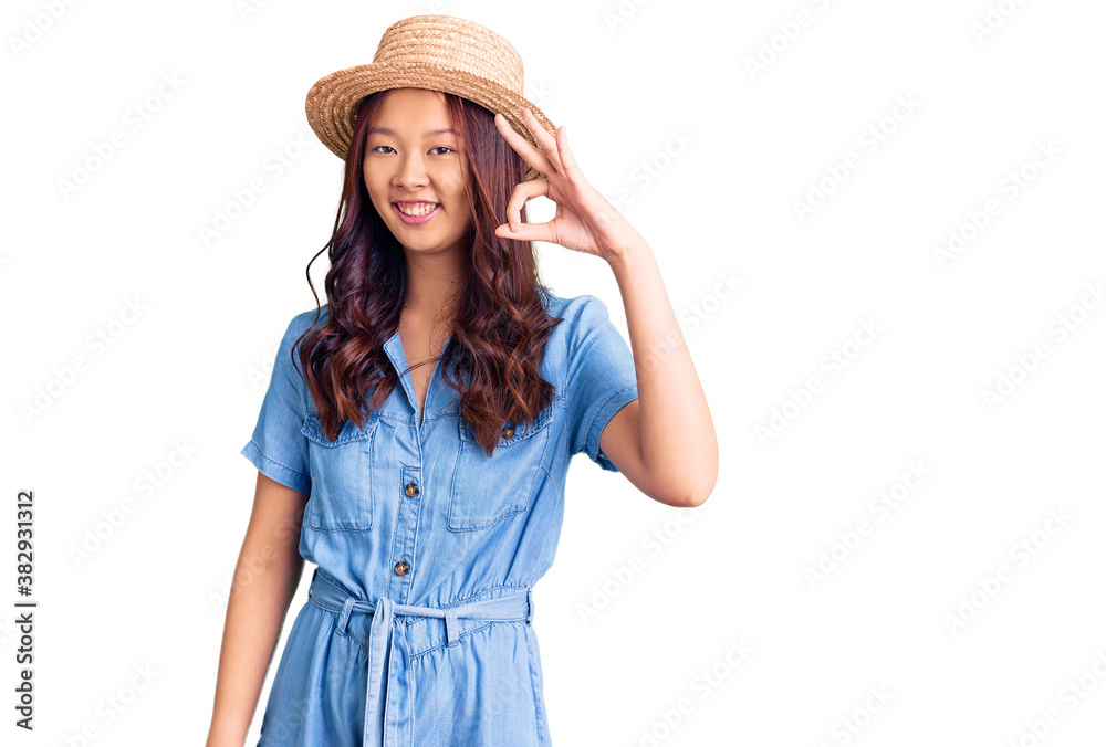 Young beautiful chinese girl wearing summer hat smiling positive doing ok sign with hand and fingers. successful expression. Young beautiful chinese girl wearing summer hat smiling positive doing ok sign with hand and fingers. successful expression.