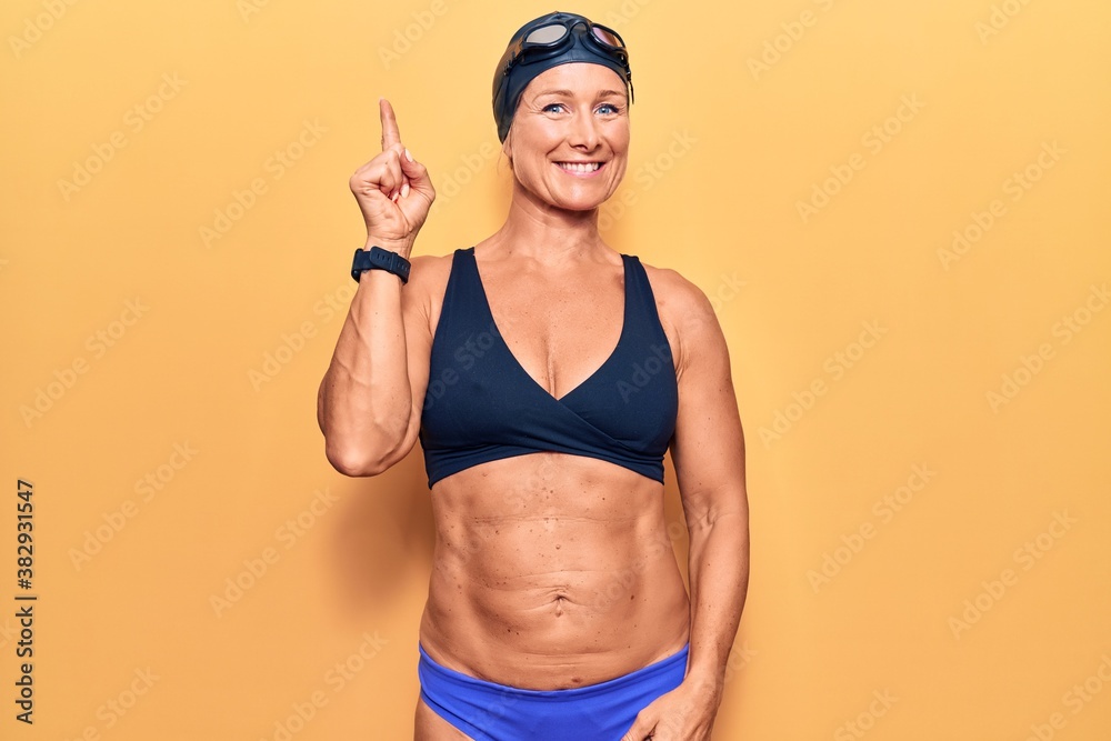 Middle age caucasian blonde woman wearing bikini,  swimming cap and diving googles pointing finger up with successful idea. exited and happy. number one.