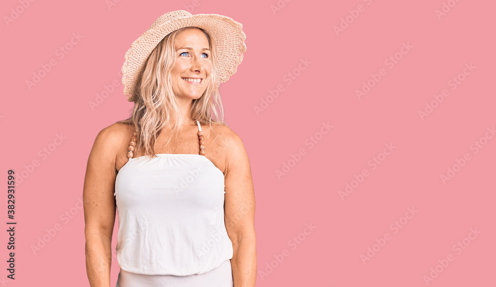 Middle age fit blonde woman wearing summer hat looking away to side with smile on face, natural expression. laughing confident. Middle age fit blonde woman wearing summer hat looking away to side with smile on face, natural expression. laughing confident.