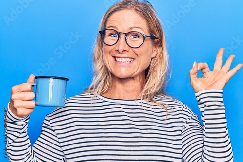 Middle age caucasian blonde woman wearing glasses drinking a cup of coffee doing ok sign with fingers, smiling friendly gesturing excellent symbol
