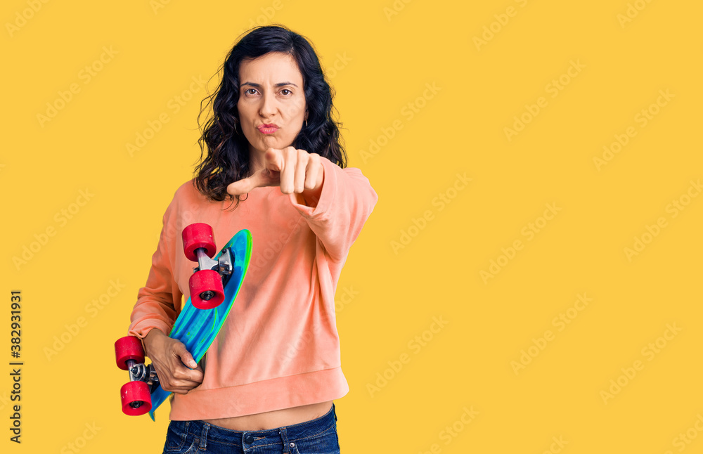Young beautiful hispanic woman holding skate pointing with finger to the camera and to you, confident gesture looking serious Young beautiful hispanic woman holding skate pointing with finger to the camera and to you, confident gesture looking serious