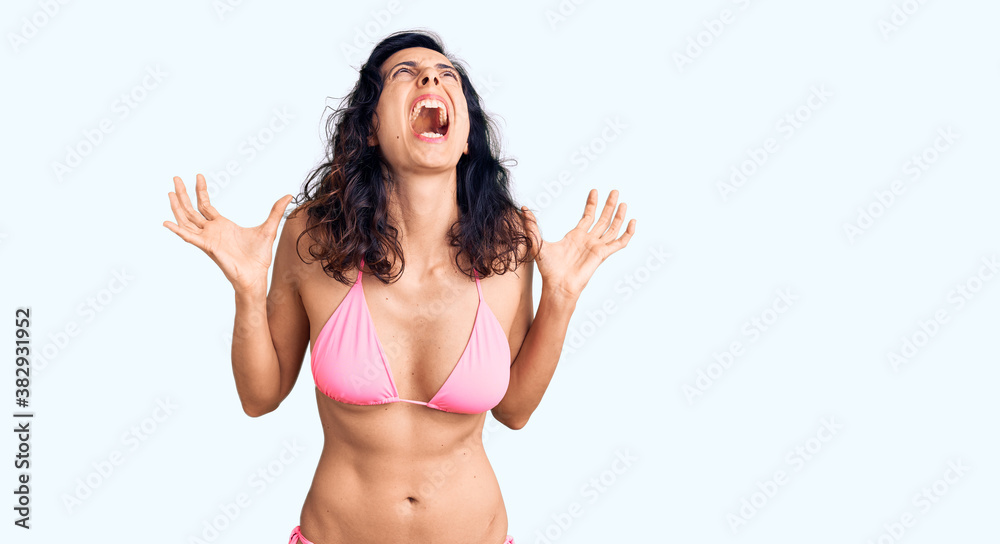 Young beautiful hispanic woman wearing bikini crazy and mad shouting and yelling with aggressive expression and arms raised. frustration concept. Young beautiful hispanic woman wearing bikini crazy and mad shouting and yelling with aggressive expression and arms raised. frustration concept.