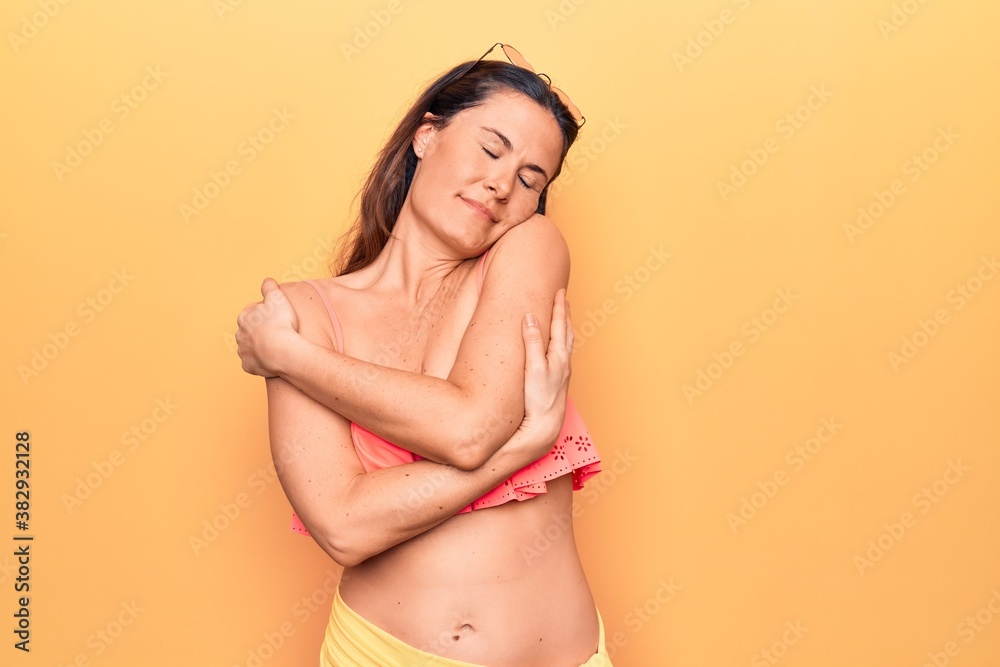 Obraz premium Young beautiful brunette woman wearing bikini standing over isolated yellow background hugging oneself happy and positive, smiling confident. Self love and self care