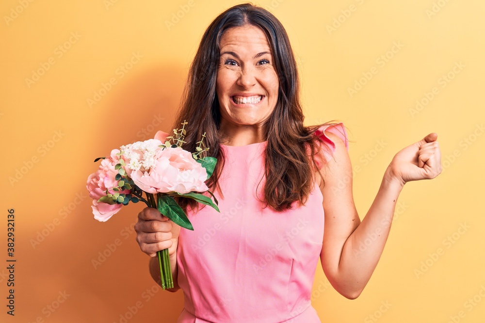 Young beautiful brunette woman holding bouquet of pink flowers over yellow background screaming proud, celebrating victory and success very excited with raised arm Young beautiful brunette woman holding bouquet of pink flowers over yellow background screaming proud, celebrating victory and success very excited with raised arm