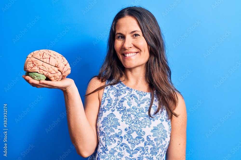 Young beautiful brunette woman asking for care memory holding brain over blue background looking positive and happy standing and smiling with a confident smile showing teeth Young beautiful brunette woman asking for care memory holding brain over blue background looking positive and happy standing and smiling with a confident smile showing teeth
