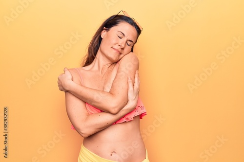 Young beautiful brunette woman wearing bikini standing over isolated yellow background hugging oneself happy and positive, smiling confident. Self love and self care