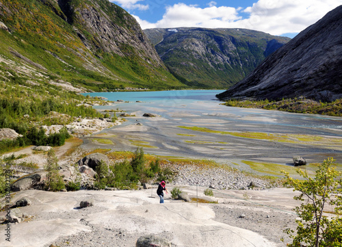 Woman Climbing Up The Rocks At Lake Nigardsbrevatnet On Her Way To The Glacier Nigardsbreen In Jostedalsbreen National Park On A Sunny Summer Day With A Clear Blue Sky And A Few Clouds
