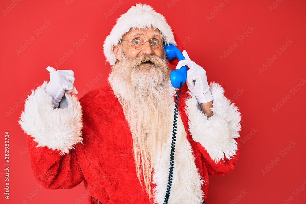 Old senior man wearing santa claus costume using vintage telephone pointing thumb up to the side smiling happy with open mouth
