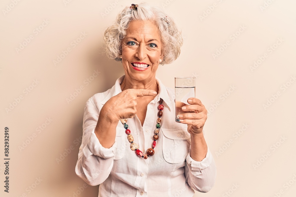 Senior grey-haired woman drinking glass of water smiling happy pointing with hand and finger Senior grey-haired woman drinking glass of water smiling happy pointing with hand and finger