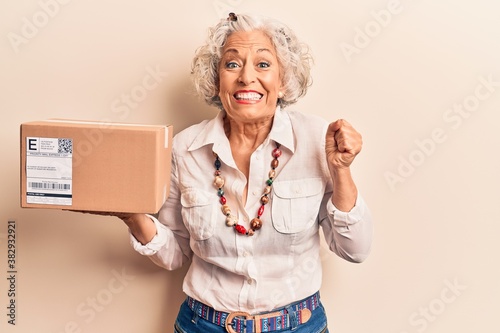 Senior grey-haired woman holding delivery package screaming proud, celebrating victory and success very excited with raised arm photo